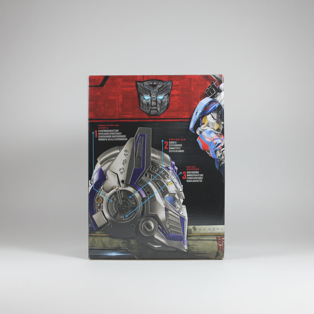 A Transformers toy box showing Optimus Prime and a detailed helmet graphic on the front, photographed standing upright on a white surface with a white backdrop and partial wooden display boards visible on both sides.