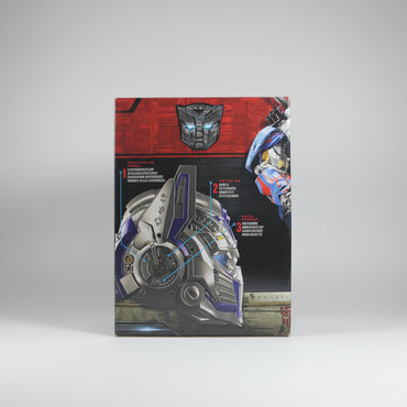 A Transformers toy box showing Optimus Prime and a detailed helmet graphic on the front, photographed standing upright on a white surface with a white backdrop and partial wooden display boards visible on both sides.